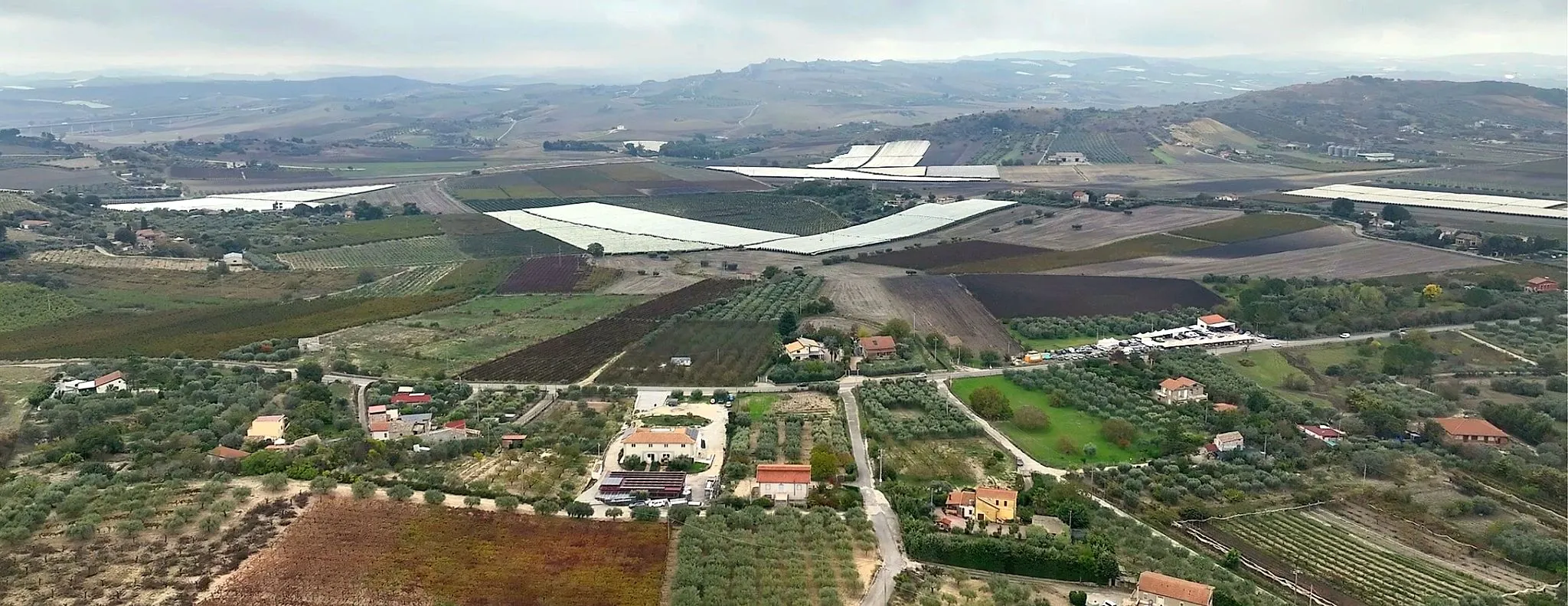 Vista aerea della campagna attorno a contrada Cusatino: colline coltivate a vigneto e uliveto, serre, case sparse e strade di campagna.